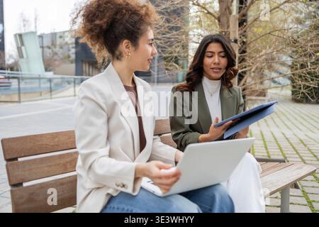 Due donne professionali sono sedute su una panchina all'aperto, impegnate in una vivace discussione. Una donna ha un notebook, mentre l'altra ha un tablet, simbo Foto Stock