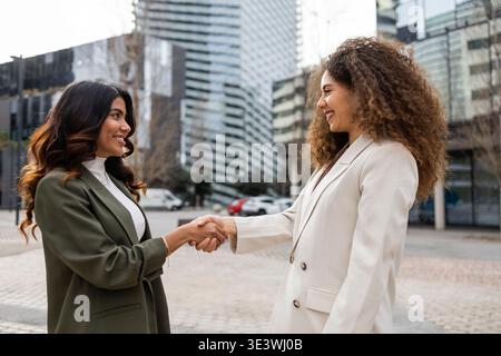 Two professional women in business attire shake hands with smiles, standing outside modern office buildings, symbolizing teamwork, success, and friend Foto Stock
