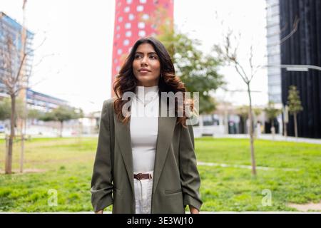 Donna d'affari sicura di sé con un blazer verde si erge all'aperto con edifici moderni sullo sfondo. Incarna determinazione e professionalità, exu Foto Stock