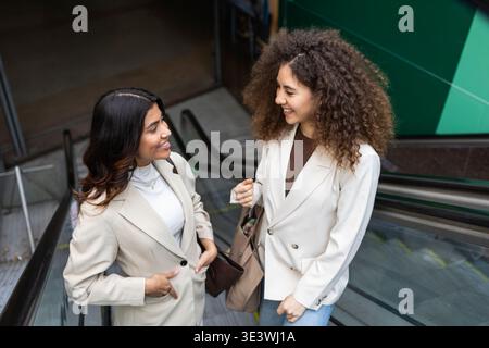 Due donne in abbigliamento da lavoro si impegnano in una conversazione vivace mentre guidano una scala mobile. Sembrano essere amici professionisti o colleghi che condividono un m Foto Stock