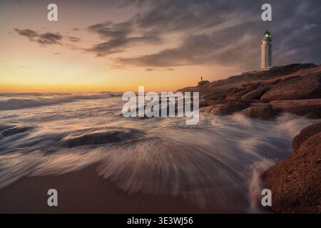 Vista mozzafiato del tramonto al faro di Trafalgar con onde che si infrangono sulla costa rocciosa, evidenziando la bellezza serena del mare e il vibrante cielo pai Foto Stock