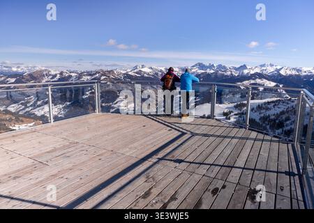 Due persone godono di una vista invernale mozzafiato da una piattaforma di osservazione in legno sulle Alpi. Le montagne innevate si estendono maestosamente sotto un blu chiaro Foto Stock
