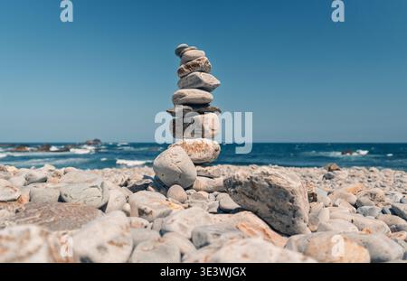 Una pila di pietre lisce e intemperie è delicatamente bilanciata sulla costa rocciosa della spiaggia di Campiechos nelle Asturie, con l'oceano blu profondo e il cielo limpido Foto Stock