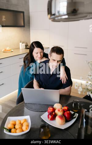 Una coppia condivide un momento a colazione nella loro cucina moderna. La donna abbraccia amorevolmente il suo partner mentre lavora su un notebook al tavolo da pranzo. Foto Stock