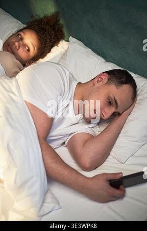 Verticale di un giovane adulto caucasico sdraiato a letto che regge lo smartphone, guardando lontano con espressione seria, mentre la giovane donna adulta nera giaceva accanto a lui sorridente e lo osservava da vicino Foto Stock