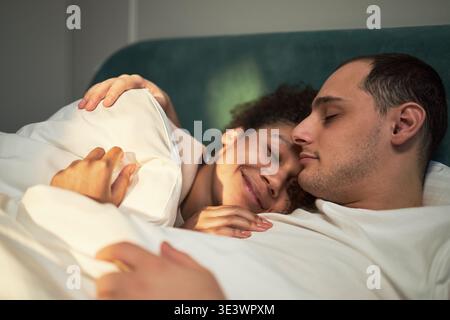 Giovane donna nera adulta e giovane uomo caucasico adulto sdraiati a letto insieme, abbracciati sotto coperta, entrambi rilassanti con gli occhi chiusi, donna sorridente dolcemente mentre coccola l'uomo Foto Stock