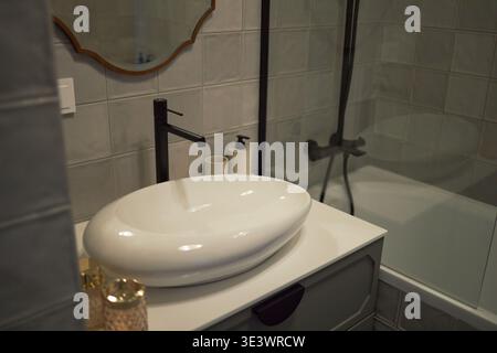 I moderni interni del bagno presentano lavandino in ceramica con rubinetto nero, dispenser di sapone sul piano, box doccia in vetro con vasca, pareti piastrellate in tonalità neutre Foto Stock