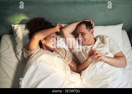 Giovane donna nera adulta e giovane uomo caucasico adulto sdraiato a letto sorridendo l'un l'altro, tenendosi per mano sotto la coperta, rilassandosi insieme in camera da letto, mostrando un'interazione affettuosa Foto Stock
