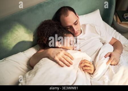 Giovane uomo adulto caucasico che abbraccia la giovane donna adulta nera mentre sdraiata a letto insieme, entrambi riposati con gli occhi chiusi, mostrando un intimo momento di coppia che si rilassa in camera da letto Foto Stock