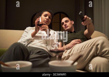 Donna nera di mezza età che parla sullo smartphone tenendo un bicchiere di vino, seduta sul divano accanto al giovane uomo caucasico che cambia canale televisivo con telecomando Foto Stock