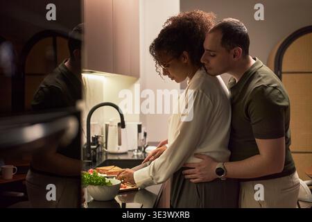 Giovane uomo caucasico adulto che abbraccia la giovane donna nera adulta da dietro mentre prepara il cibo al bancone della cucina, entrambi vicini, uomo che riposa delicatamente la testa sulla spalla Foto Stock