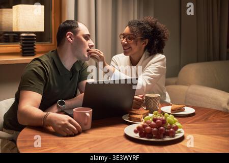 Giovane uomo caucasico adulto seduto a tavola con la giovane donna nera adulta che gli dà da mangiare, sorridente e interagente, laptop e piatti di frutta e panini sul tavolo Foto Stock