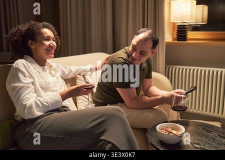 Uomo caucasico e donna nera seduti sul divano bevendo vino rosso e parlando, uomo con telecomando e donna sorridente, ciotola di spuntini sul tavolo di fronte a loro, ambiente interno serale Foto Stock