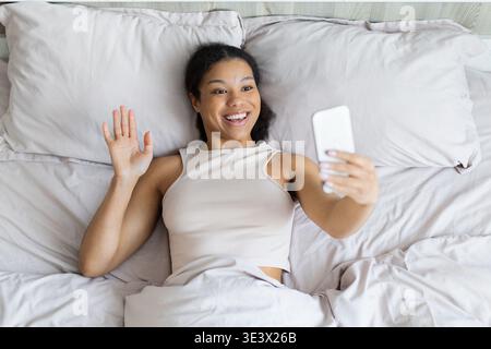 Giovane donna nera sdraiata a letto sorridente e ondeggiante mentre tiene in mano lo smartphone Foto Stock