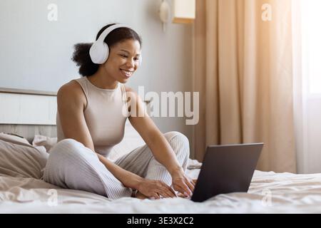 Giovane donna nera con le cuffie sedute sul letto utilizzando comodamente il computer portatile Foto Stock