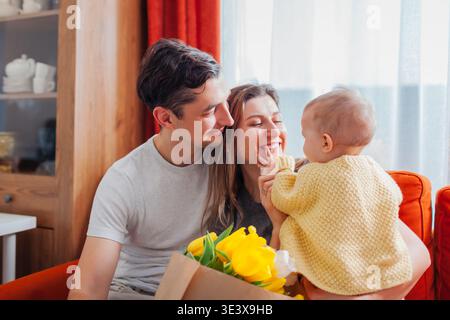 Famiglia felice che festeggia la festa della mamma. L'uomo ha donato alla moglie un bouquet di fiori a casa. I genitori giocano con la bambina. Foto Stock