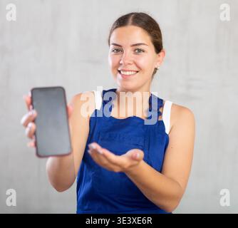 Felice lavoratrice che indossa abiti da lavoro che mostrano lo schermo del suo smartphone Foto Stock