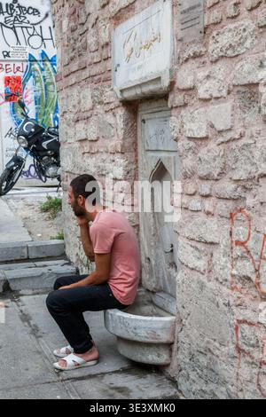 L'uomo barbuto in t-shirt rosa si siede su una nicchia decorata di fontane in marmo ottomano, parlando al telefono in una strada posteriore di Istanbul fiancheggiata da graffiti Foto Stock
