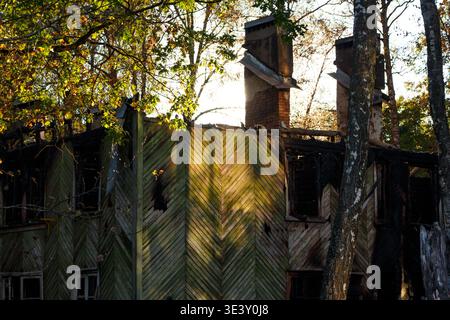 I resti carbonizzati di una vecchia casa in legno sono stati abbandonati, con la luce autunnale che si diffonde tra gli alberi ricoperti. Una scena toccante di degrado e abbandono Foto Stock