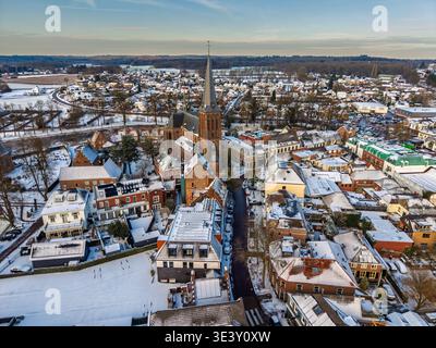 Vista aerea di un pittoresco villaggio europeo coperto di neve al tramonto, caratterizzato da una storica chiesa in mattoni con un'alta guglia, tradizionali tetti di tegole rosse Foto Stock