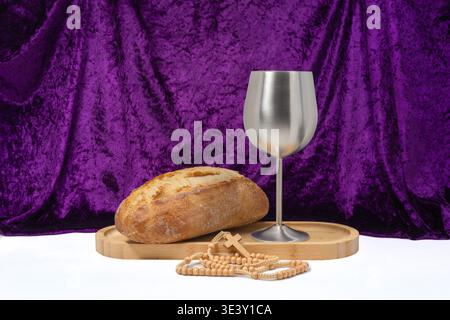 Pane con calice e rosario su un vassoio di legno su un ricco fondo in tessuto viola. Rappresenta la Quaresima, la preghiera e la comunione cristiana simbolis Foto Stock
