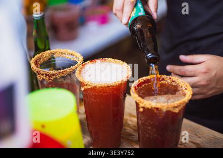 Primo piano di un barista che versa birra scura da una bottiglia in bicchieri michelada preparati con bordi in polvere di sesamo e peperoncino. Foto Stock