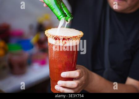 Persona che versa due birre contemporaneamente in un bicchiere michelada preparato con peperoncino, semi di sesamo e bordo di salsa camoscio. Foto Stock