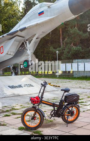 Una robusta FAT bike elettrica pieghevole con cerchi arancioni vibranti si distingue per essere pronta all'avventura. Il maestoso jet MIG-29 sullo sfondo aggiunge un tocco di alta qualità Foto Stock