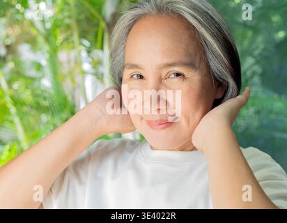 Primo piano di una donna asiatica matura felice con i capelli grigi che si toccano a casa. Ritratto, concetto di stile di vita pensionato. Foto Stock