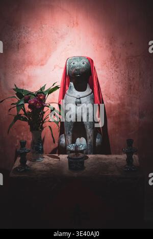 Statua del cane guardiano di pietra drappeggiata in tessuto rosso su un altare del santuario con incenso bruciatore e fiori, Ponte coperto giapponese, Hoi An, Vietnam. Foto Stock