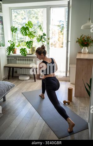Donna positiva matura che si allena a casa, facendo esercizi di stretching sul tappetino in salotto. Foto Stock