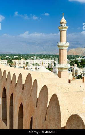 Il forte di Nizwa mostra una vista della moschea e della città sottostante. Il sole splende sugli edifici con montagne sullo sfondo. Foto Stock