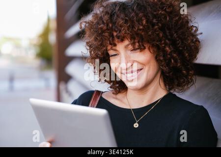 Donna sorridente con capelli ricci e maglione nero si impegna con il suo tablet all'aperto sotto il sole Foto Stock