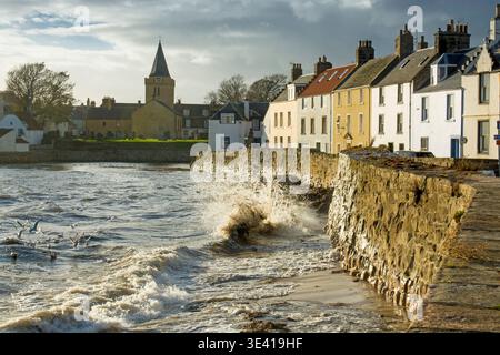 Le onde tempestose si infrangono contro il muro di pietra del mare di Anstruther, Fife, Scozia, illuminando il villaggio sul mare, il campanile della chiesa e le file storiche di Hou Foto Stock