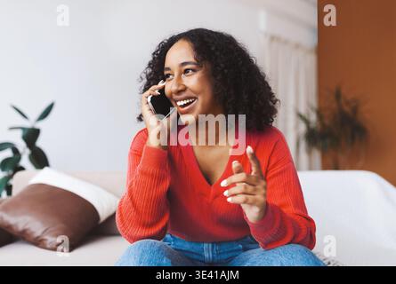 Una donna felice parla al telefono mentre si siede sul divano nell'accogliente spazio abitativo Foto Stock