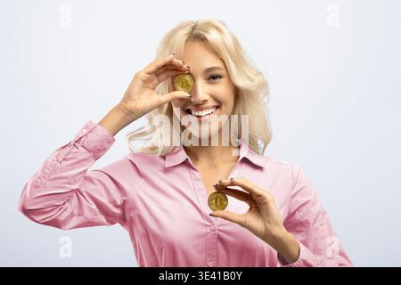 Donna tiene due monete Bitcoin mentre sorride e guarda la fotocamera in un ambiente leggero Foto Stock