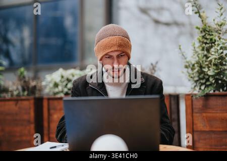 Giovane uomo che lavora su un computer portatile in un bar all'aperto con berretto e giacca invernale Foto Stock
