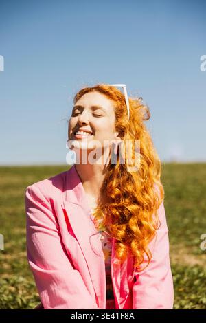 Felice donna dai capelli di zenzero con gli occhi chiusi che prende il sole in un campo verde Foto Stock