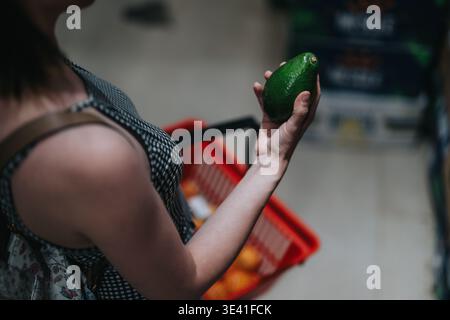 Donna che tiene in mano un avocado mentre fa la spesa con un cestino rosso in un supermercato Foto Stock
