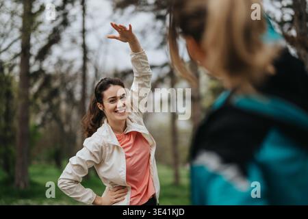 Una donna in un parco sorride e alza la mano durante un allenamento all'aperto con un amico nelle vicinanze Foto Stock
