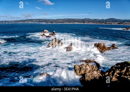 Rocce aspre incontrano onde che si infrangono sotto il cielo azzurro. La potenza oceanica sfreccia contro la pietra frastagliata con schiuma bianca. Colline lontane fanno da cornice all'orizzonte Foto Stock