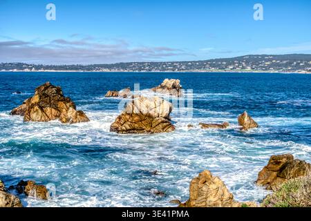 Le onde turchesi si schiantano contro le aspre rocce marroni. Il cielo si illumina di blu brillante, punteggiato da morbide nuvole. Colline lontane incorniciano la costa calma e soleggiata. Roccia Foto Stock