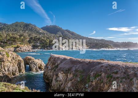 Le scogliere illuminate dal sole incorniciano le onde turchesi dell'oceano. Le nuvole bianche si estendono sopra le colline boscose. Gli archi in pietra robusti resistono contro le onde che si infrangono. Luminoso b Foto Stock