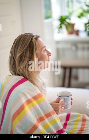 Donna sorridente e premurosa che si rilassa a letto con una tazza di caffè comodamente seduta avvolta in coperta. Foto Stock