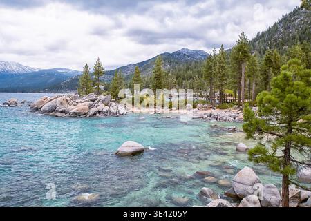 Le acque cristalline del lago illuminano il turchese vicino alla costa rocciosa. I pini fiancheggiano il bordo, alti e verdi. Montagne innevate si innalzano dietro una fitta foresta. Nuvola di cielo Foto Stock