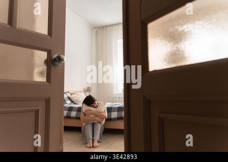 Donna che abbraccia il cuscino mentre siede sul pavimento della camera da letto, si sente sola, triste e soffre di problemi di salute mentale Foto Stock