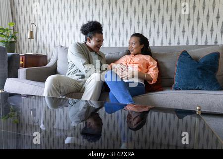 Coppia seduta su divano grigio, tavolo di vetro che riflette entrambi, uomo che mette mano su donna asiatica incinta Foto Stock
