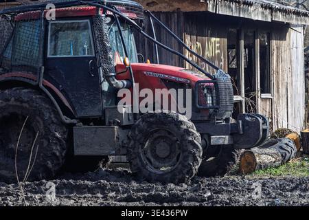 Vinkovci, Croazia - 27 febbraio 2026: Un trattore fangoso rosso si trova accanto a un capannone di legno intemprato e taglia tronchi dopo lavori di silvicoltura e di legname su un terreno umido Foto Stock