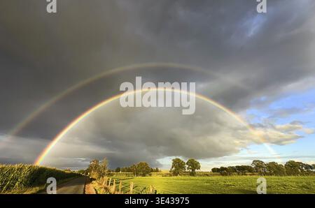 Uno splendido doppio arcobaleno si arrocca elegantemente su campi verdi e vivaci sotto le nuvole scure che incombono Foto Stock