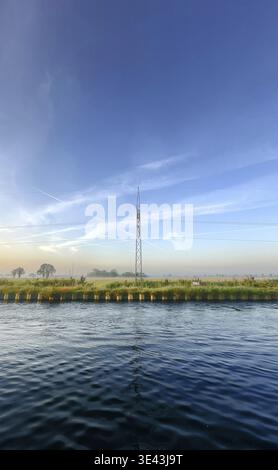 Una tranquilla scena fluviale che mostra un'alta torre di comunicazione adagiata su un cielo azzurro e lussureggiante vegetazione tutt'intorno Foto Stock